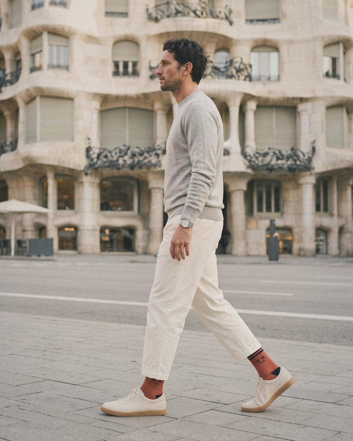 A man in a grey knit and cream trousers walks past Gaudí's Casa Milà in Barcelona, wearing the MUNT Blood sock
